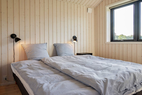 Soveværelse med dobbeltseng, træpaneler, to væglamper og vindue i feriebolig for 8 i Aakirkeby, Danmark.
