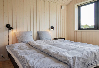 Dormitorio con cama doble, paredes de paneles de madera, dos apliques y ventana en casa vacacional en Aakirkeby, Dinamarca.