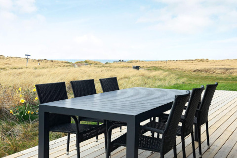 Outdoor patio with black dining table and six chairs, overlooking dunes and coastline in Hjørring, Denmark.
