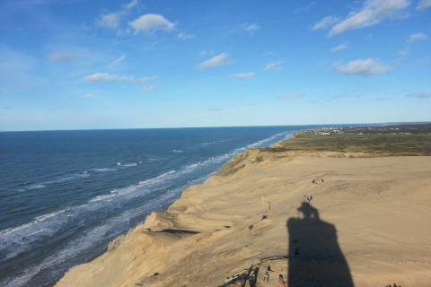 Zicht op de kustlijn en duinen nabij Hjørring, Denemarken, onder heldere blauwe lucht met vuurtorenschaduw.