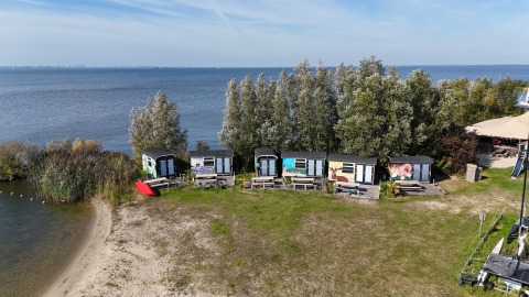 Luftfoto af Tiny Wagon Lodge med små farverige hytter ved søbredden og grønne træer i baggrunden.
