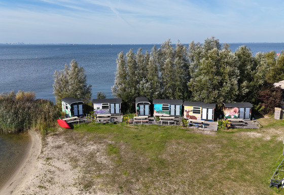 Luftfoto af Tiny Wagon Lodge med små farverige hytter ved søbredden og grønne træer i baggrunden.