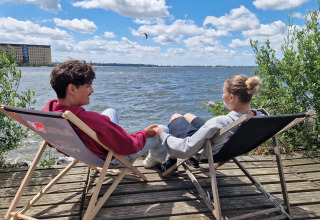 To personer sidder afslappet i liggestole ved vandet ved Tiny Wagon på Marina Parcs Almere, Holland.