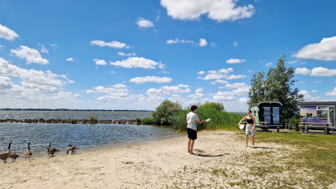 Deux personnes jouent sur la plage près du Tiny Wagon à Marina Parcs Almere, Pays-Bas, avec des oies devant.