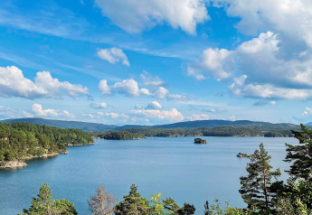 Vista di un lago tranquillo e colline verdi dalla casa vacanze per 4 persone a Reksteren sotto un cielo azzurro.