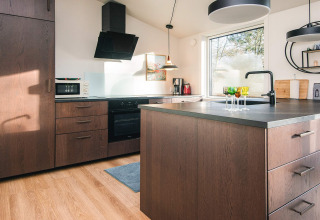 Moderne keuken met houten kasten, kookeiland, groot raam en veel licht in een vakantiehuis in Ans By, Denemarken.