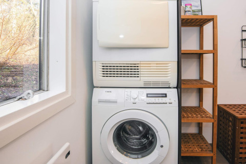 Waskamer in het vakantiehuis met wasmachine, droogkast, houten rekken en zicht op de tuin door het raam.