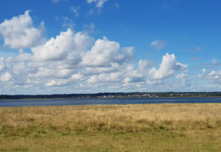 Vue depuis la maison de vacances 10 personnes à Hadsund, Danemark, sur champ, eau et ciel bleu nuageux.