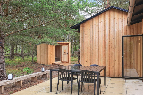 Patio exterior de una casa vacacional en Ålbæk, Dinamarca, con mesa, sillas y sauna en un bosque.