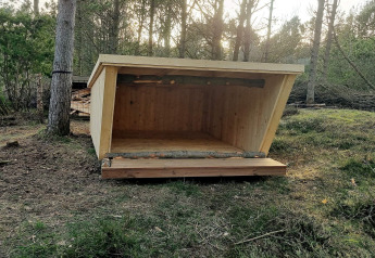 Houten schuilhut in het bos bij 8-persoons vakantiehuis in Ålbæk, Denemarken, omringd door bomen.