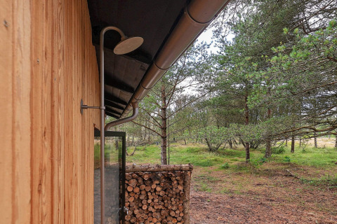 Outdoor shower attached to a wood-paneled lodge with firewood stack, surrounded by woods in Ålbæk, Denmark.