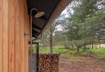 Ducha exterior en una cabaña con paneles de madera y leña amontonada, rodeada de bosque en Ålbæk, Dinamarca.