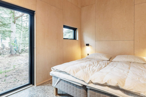 Dormitorio moderno en casa vacacional en Ålbæk, Dinamarca, con paredes de madera y grandes ventanas al bosque.