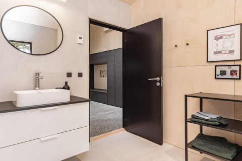Modern bathroom with black door, white sink, round mirror, and shelving in a holiday home in Ålbæk, Denmark.