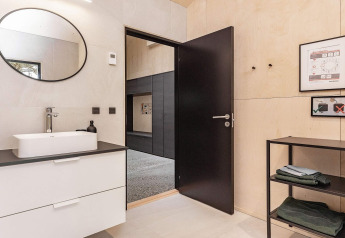 Modern bathroom with black door, white sink, round mirror, and shelving in a holiday home in Ålbæk, Denmark.