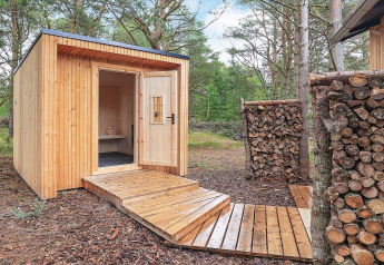 Sauna de madera con terraza y pilas de leña, situada en un bosque en Ålbæk, Dinamarca.