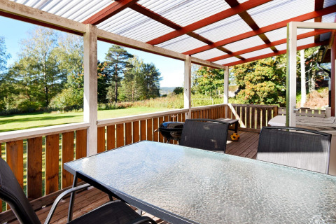 Overdækket træterrasse med glasbord og stole, grill og udsigt til grøn have ved chalet i Sunnemo, Sverige.