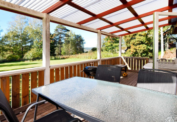Overdækket træterrasse med glasbord og stole, grill og udsigt til grøn have ved chalet i Sunnemo, Sverige.