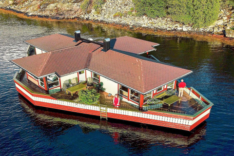 Woonboot 38508-FISKHOLMEN drijft aan de oever, met twee mensen aanwezig op het terras van het dek.