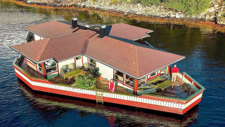 Hus på vandet, navngivet 38508-FISKHOLMEN, flyder ved kysten med to personer på dækket.