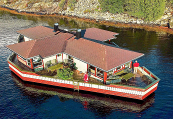 Woonboot 38508-FISKHOLMEN drijft aan de oever, met twee mensen aanwezig op het terras van het dek.