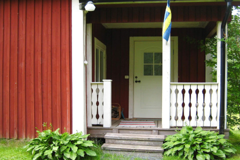 Entrada de una cabaña con bandera sueca, paneles de madera rojos y blancos y plantas verdes en Sunnemo, Suecia.