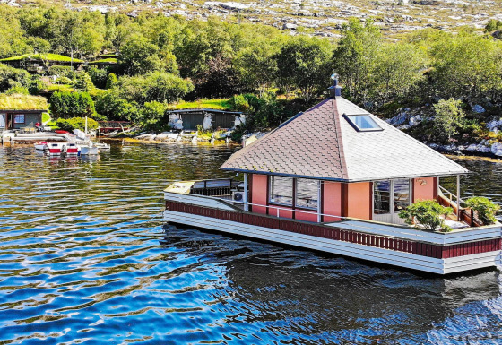 Houseboat 38507-SJØSTJERNEN floating on a lake, surrounded by trees and hills on a bright sunny day.