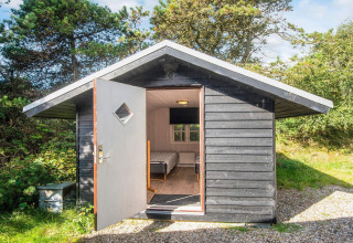 Zwart houten huisje met open deur, bed zichtbaar, omgeven door bomen in Ho Bay, Denemarken.