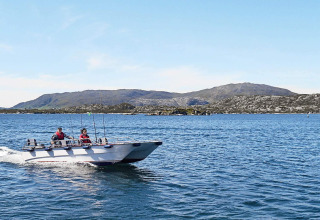 Dos personas navegan en una lancha cerca de Fiskholmen, Dinamarca, con montañas al fondo.