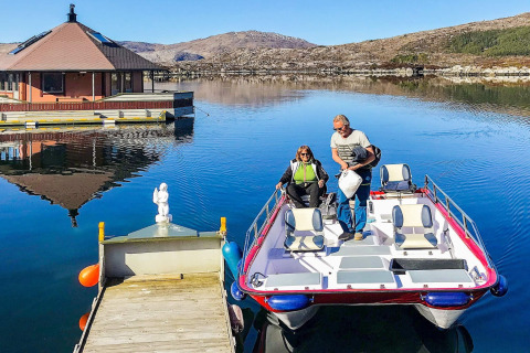 Two people arrive at the houseboat named 38507-SJØSTJERNEN on a calm lake surrounded by hills in Denmark.