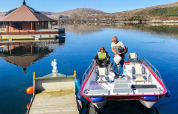 Two people arrive at the houseboat named 38507-SJØSTJERNEN on a calm lake surrounded by hills in Denmark.
