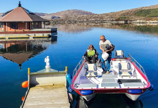 Two people arrive at the houseboat named 38507-SJØSTJERNEN on a calm lake surrounded by hills in Denmark.