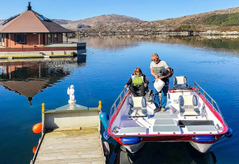 Two people arrive at the houseboat named 38507-SJØSTJERNEN on a calm lake surrounded by hills in Denmark.