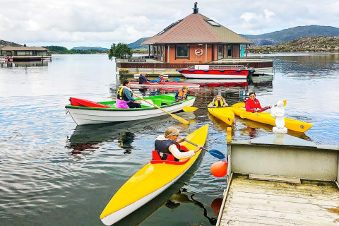 People paddling kayaks and boats near the houseboat '38507-SJØSTJERNEN' on a lake in Denmark, with hills.