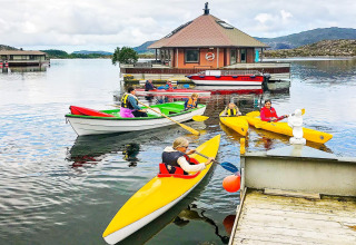 People paddling kayaks and boats near the houseboat '38507-SJØSTJERNEN' on a lake in Denmark, with hills.