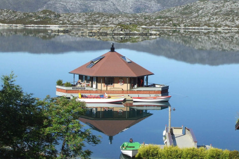 Houseboat «38507-SJØSTJERNEN» floats peacefully on a calm lake in Denmark with mountains in the background.