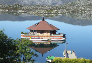 Houseboat «38507-SJØSTJERNEN» floats peacefully on a calm lake in Denmark with mountains in the background.