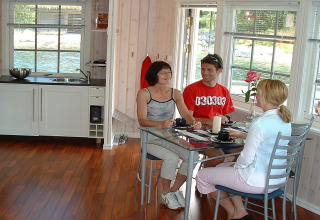 Three people sit at a glass table in the bright kitchen of houseboat 38507-SJØSTJERNEN in Denmark.