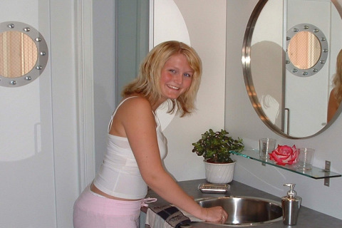 Woman smiling at sink in modern bathroom on houseboat ‘38507-SJØSTJERNEN’ in Denmark, with mirror and plant.