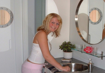 Woman smiling at sink in modern bathroom on houseboat ‘38507-SJØSTJERNEN’ in Denmark, with mirror and plant.