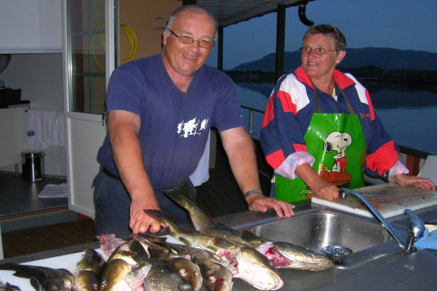 Two people cleaning fish at a sink on the houseboat «38507-SJØSTJERNEN» in Denmark with a scenic view.