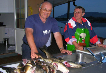 Two people cleaning fish at a sink on the houseboat «38507-SJØSTJERNEN» in Denmark with a scenic view.