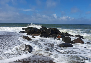 Waves crash against rocks on the beach near an 8-person holiday home in Ulfborg, Denmark, blue sky above.