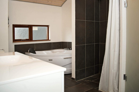 Moderne badeværelse med hjørnebadekar, bruseniche med mørke fliser og vindue i feriehus i Ulfborg, Danmark.