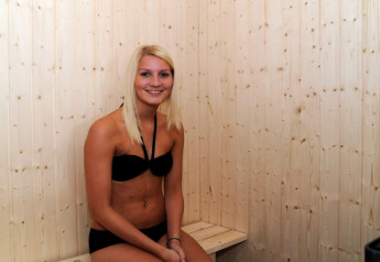Ung kvinde i sort bikini sidder og smiler på bænk i en træsauna i feriehytten til 8 personer i Ulfborg, Danmark.