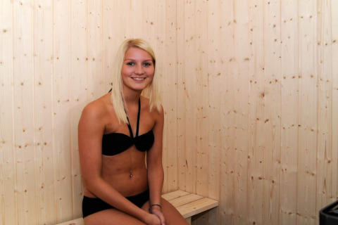 Ung kvinde i sort bikini sidder og smiler på bænk i en træsauna i feriehytten til 8 personer i Ulfborg, Danmark.