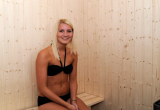Joven con bikini negro sonríe sentada en una sauna de madera en casa de vacaciones para 8 personas en Ulfborg, Dinamarca.