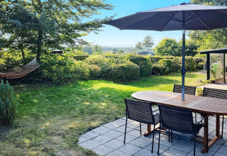 Giardino con tavolo in legno, sedie nere, ombrellone e amaca alla casa vacanza per 4 persone ad Allingåbro, Danimarca.