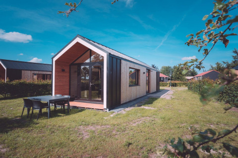 Moderne Glamping-Unterkunft mit Panoramafenstern und Gartenmöbeln auf grünem Rasen unter blauem Himmel.