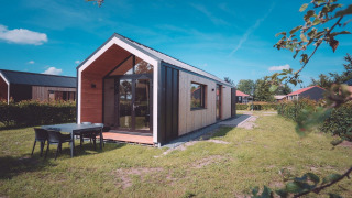 Cabaña glamping moderna con grandes ventanas y mesa exterior, rodeada de césped verde y cielo azul claro.